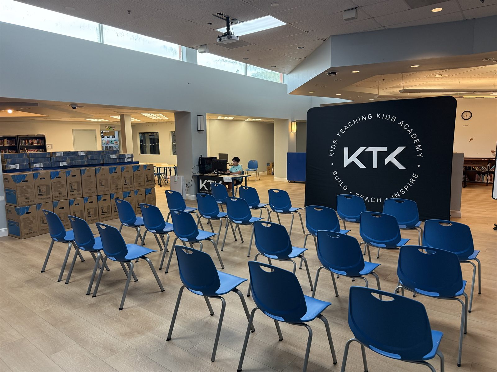 An empty room with rows of blue chairs and a wall of stacked computer case boxes before camp starts