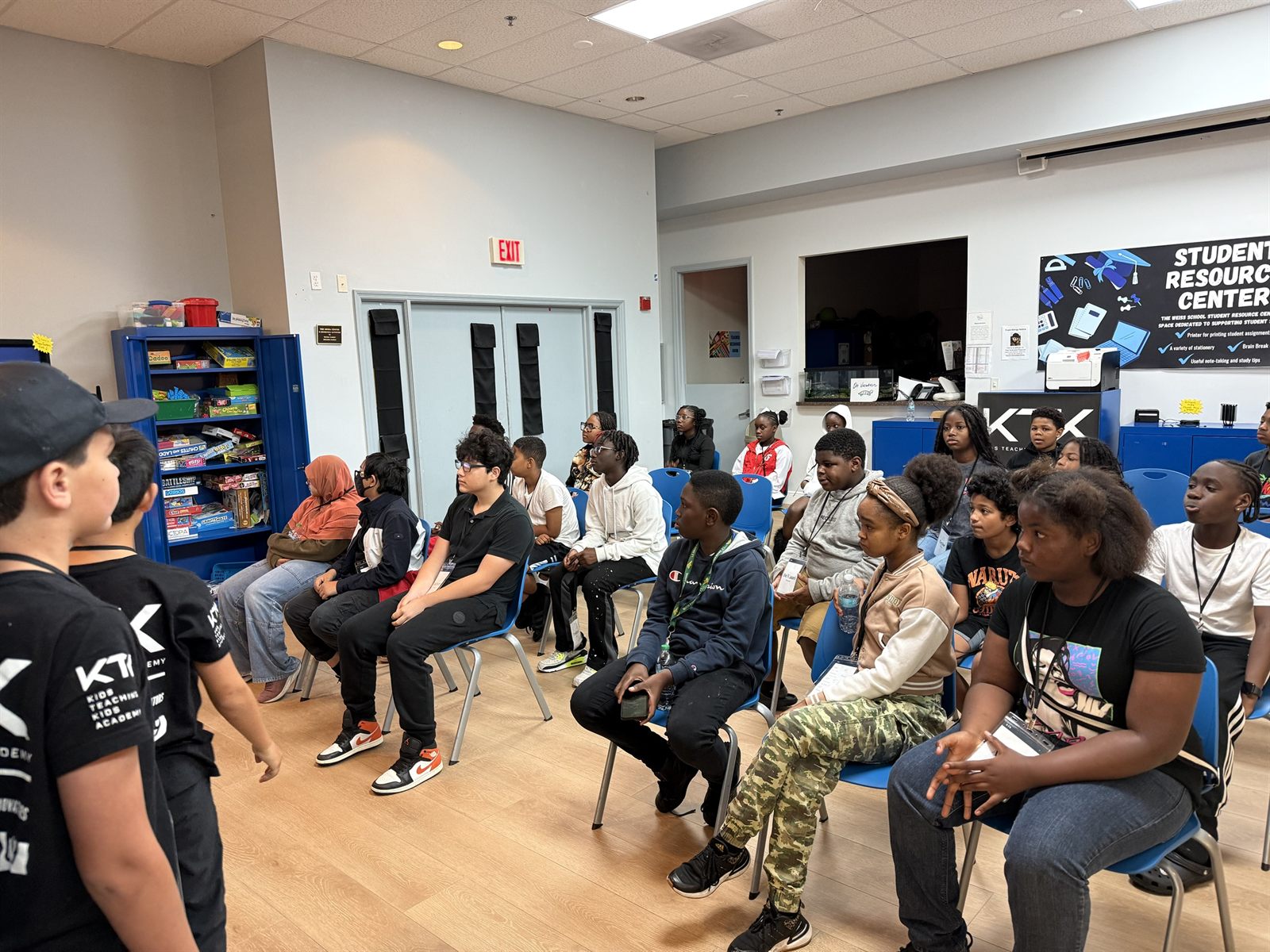 A room of campers seated in blue chairs, listening to teachers at the front