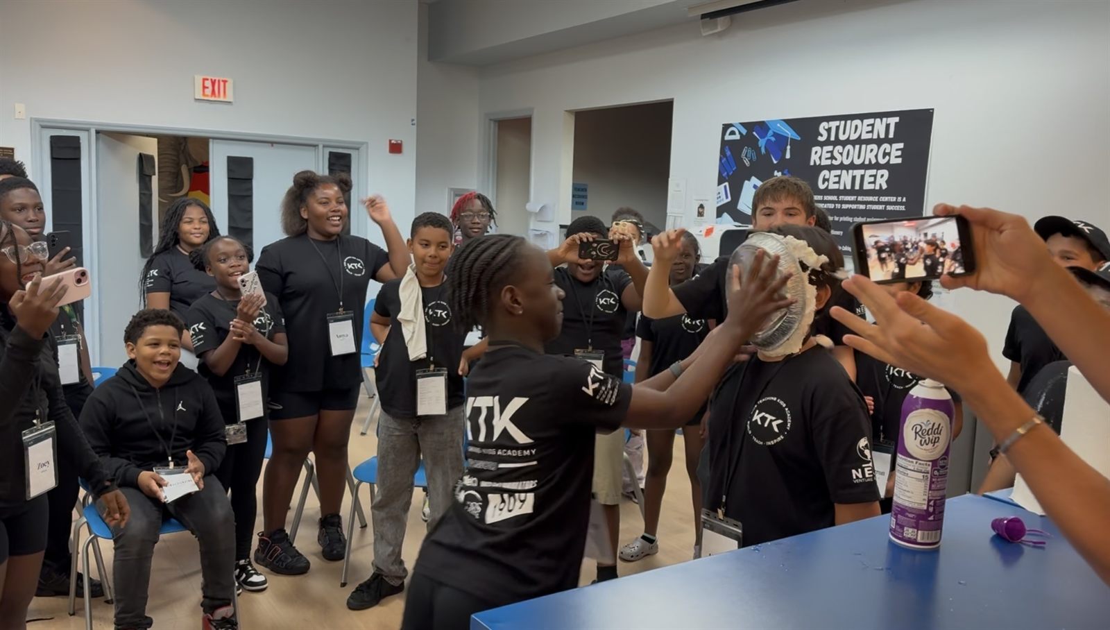 A camper pushing a whipped-cream pie into a teacher's face while the room cheers