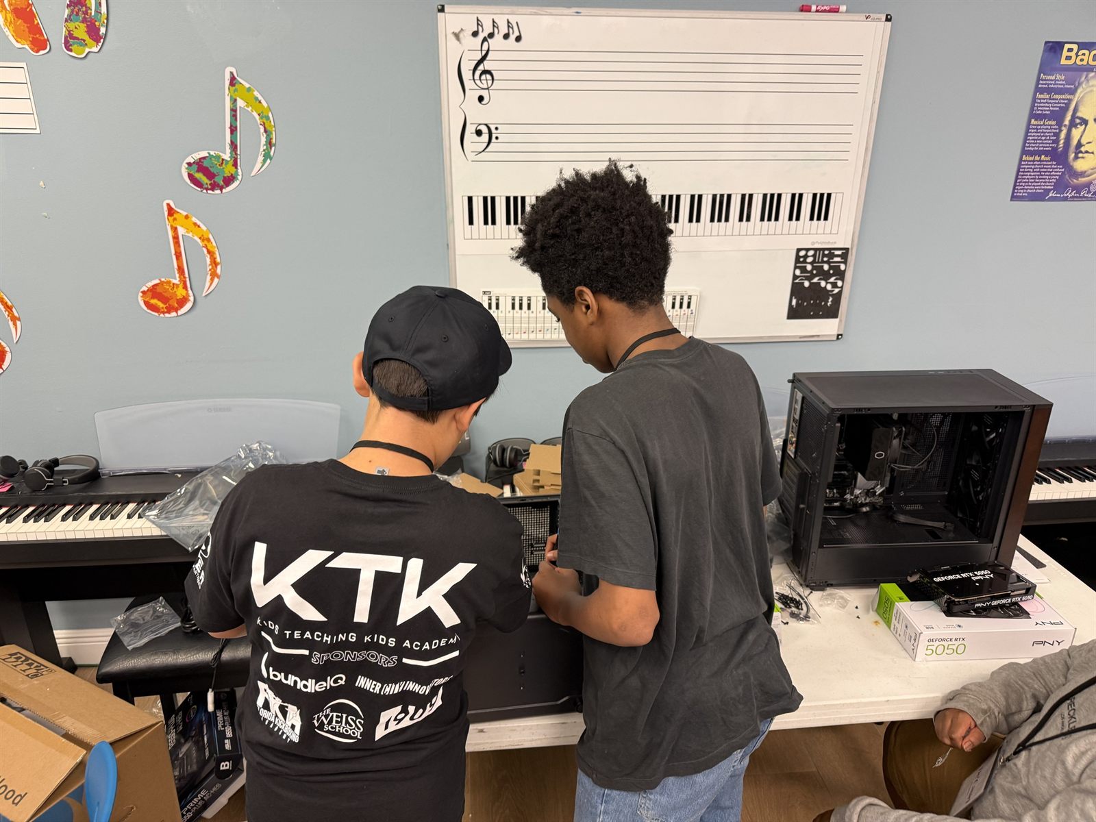 The back of a teacher's black KTK shirt listing the sponsors, in a music classroom