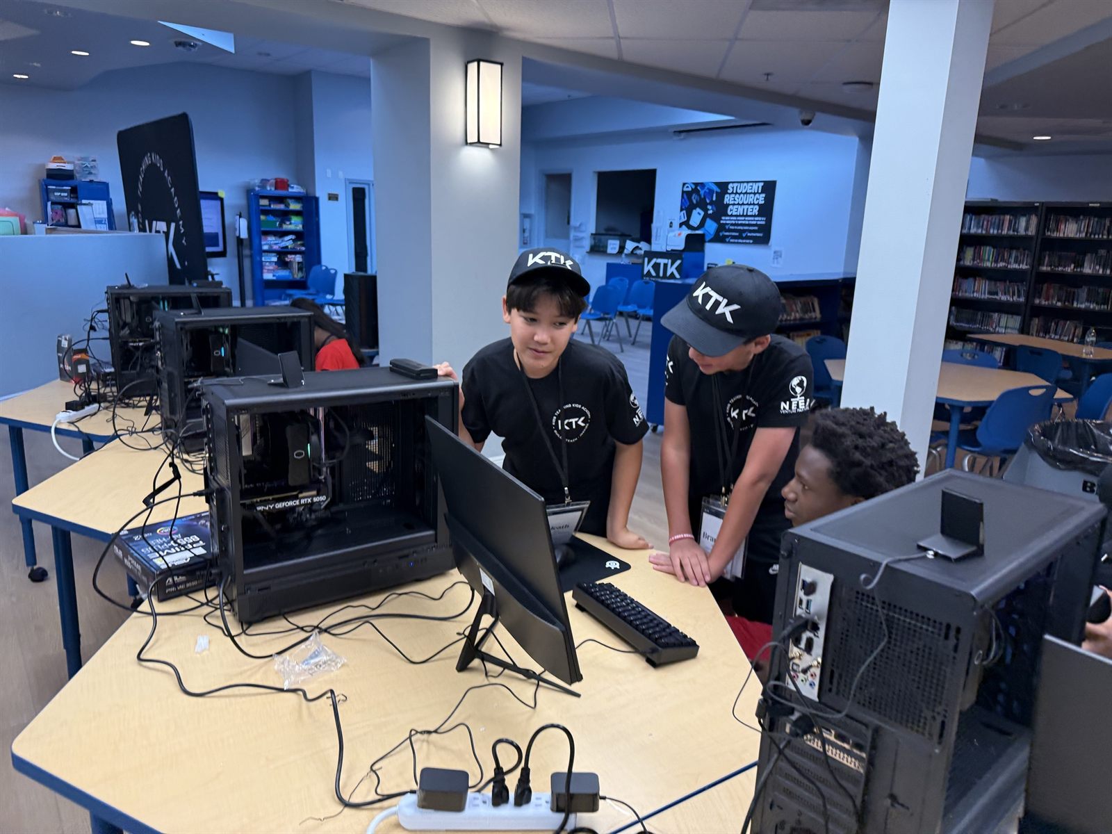 Two teachers in KTK caps checking a build at an open case with the graphics card showing