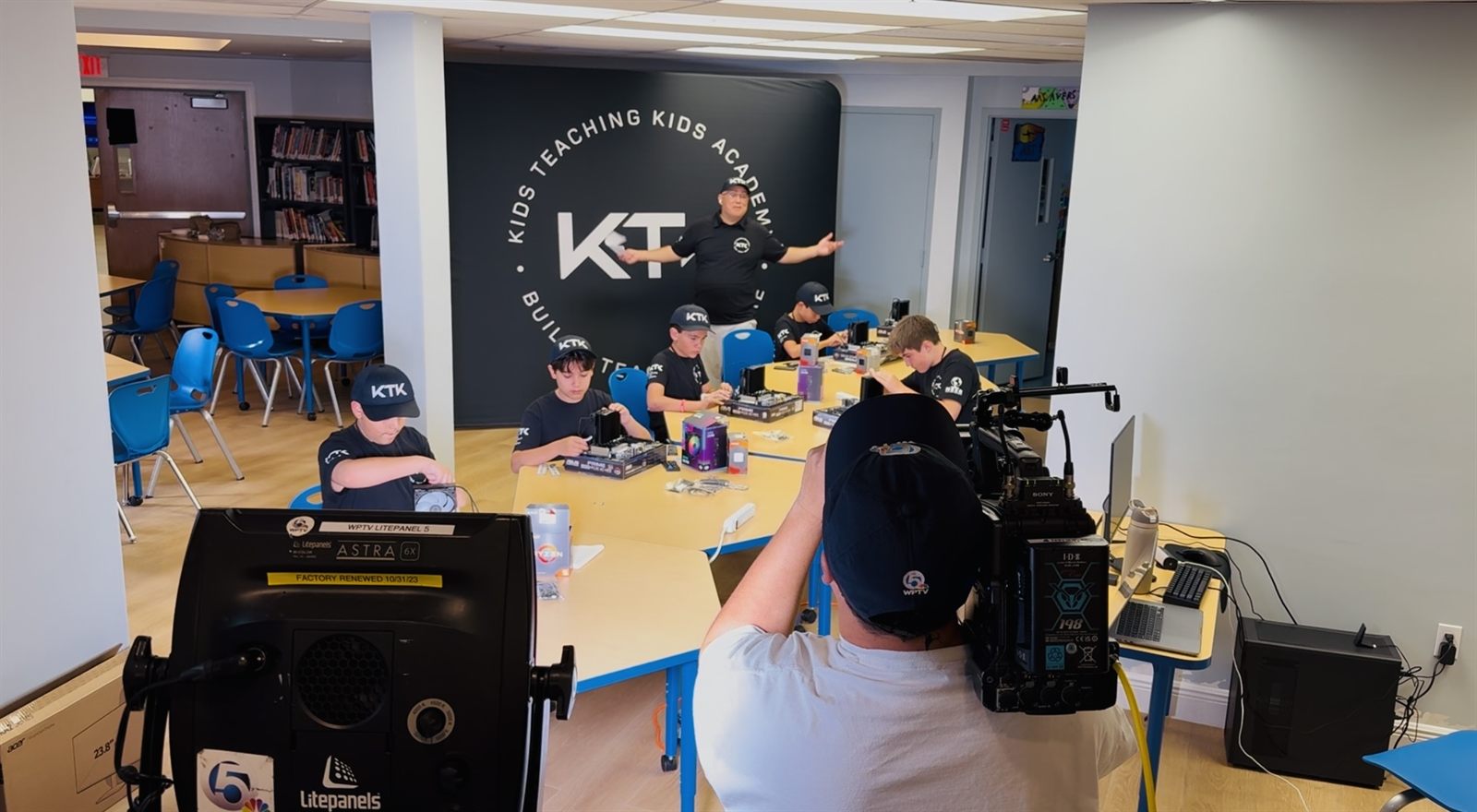 A WPTV camera operator filming teachers building at a table in front of the KTK backdrop
